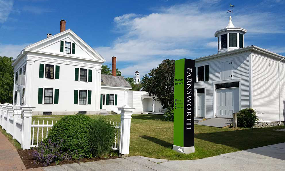 The Farnsworth Homestead on the left with the Homestead's Carriage House on the right. In between is a tall column-shaped sign for the Farnsworth in green and black.